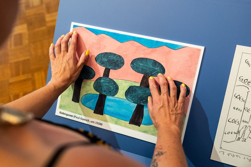 Auf einem Tisch eine taktile Reproduktio neines Gemäldes von Paul Goesch. Es zeigt einen See vor Gebirgslandschaft, umgeben von Bäumen. Hände betasten das Bild.