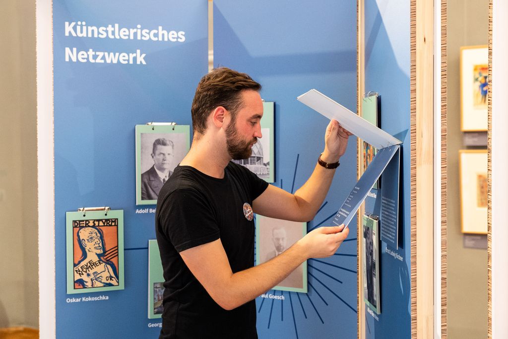 Ein bärtiger Junger Mann steht vor einer Ausstellungstafel. Einzelne Elemente auf der Tafel sind klappbar. Der Mann liest eine dier Klapptafeln.