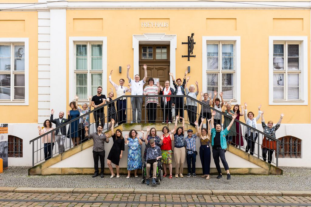 Eine barocke Hausfassade. Davor, auf einer Treppe gruppiert, eine Gruppe von ca. 20 Menschen. Sie lachen und werfen die Arme in die Luft